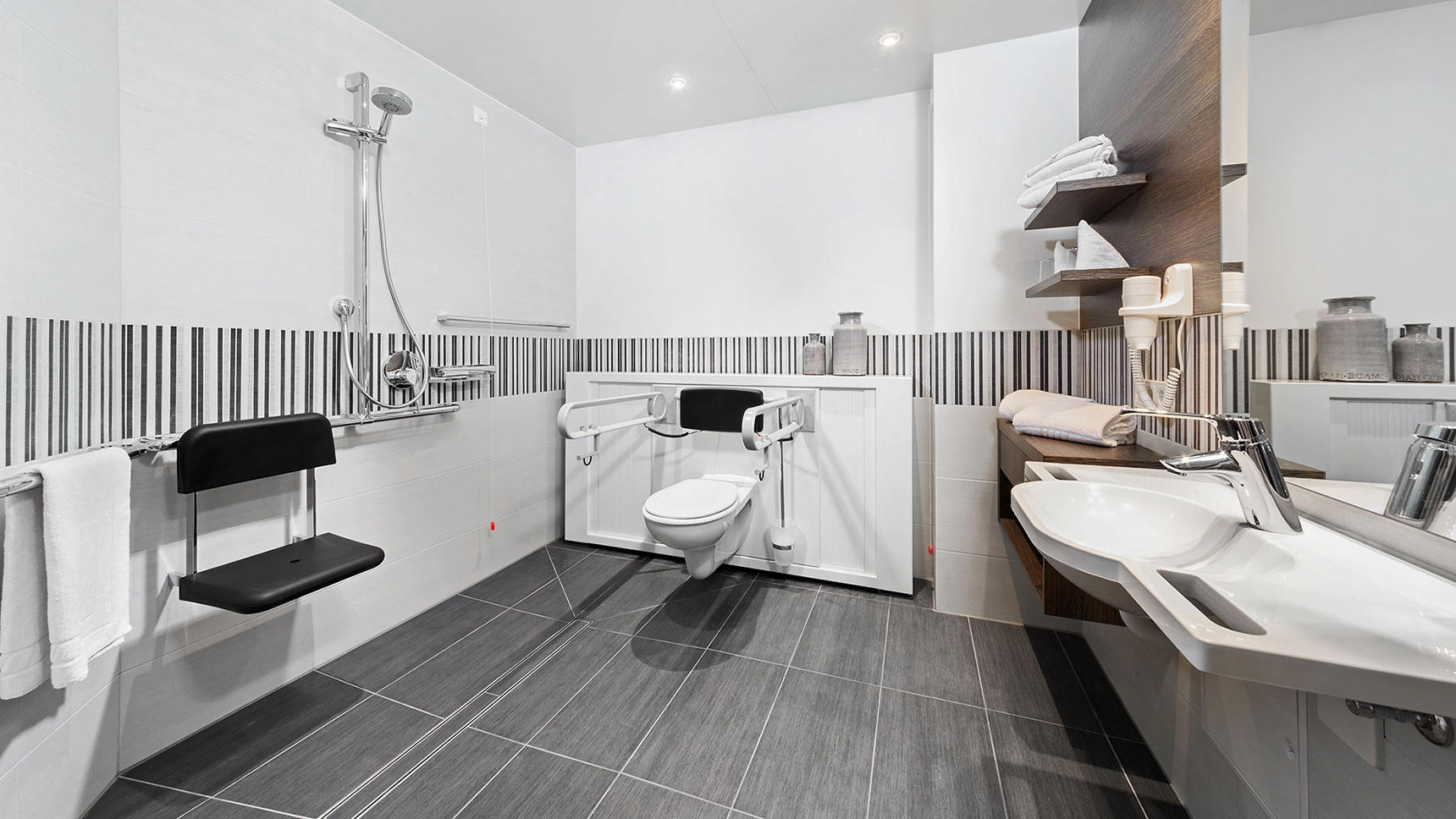 Ein großräumiges Badezimmer mit offener Dusche, Sitzplatz und Toilette mit Haltegriffen.