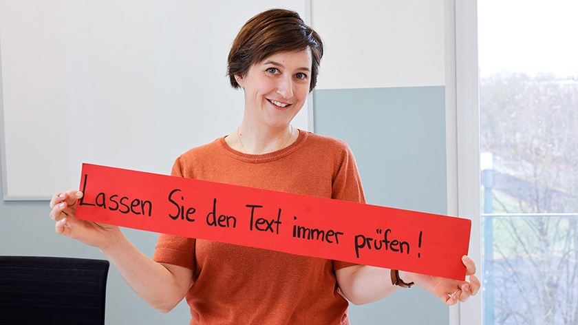 Carola Nagel hält ein Papierbanner in der Hand mit der Aufschrift: „Lassen Sie den Text immer prüfen!“.