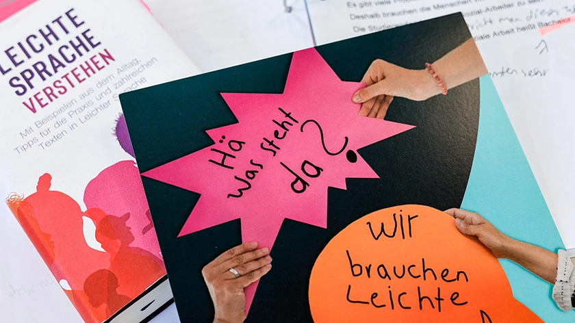 Mehrere Printmaterialien liegen ausgebreitet da, unter anderem ein Buch über Leichte Sprache und eine Collage mit Sprechblasen, in denen steht: „Hä was steht da?“ und „Wir brauchen Leichte Sprache“. 