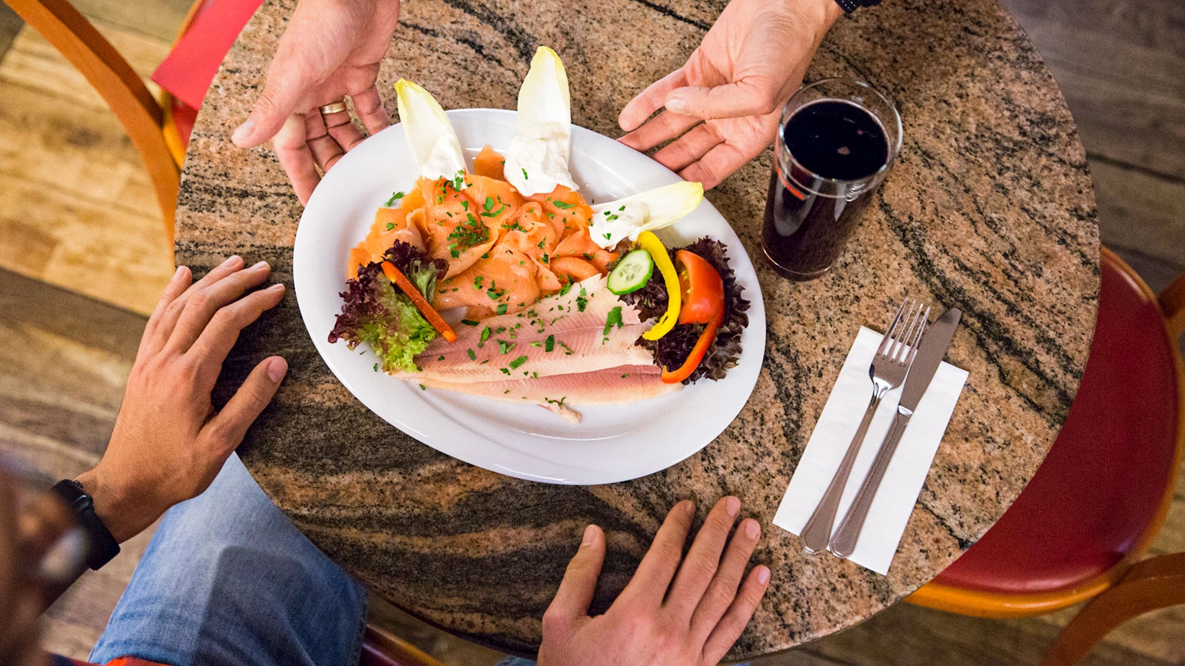 Eine Person stellt einen kunstvoll garnierten Teller mit Fisch und Gemüse auf einem Tisch ab. Zu sehen sind nur ihre Hände, sowie die Hände der Person, die das Essen erhält. 