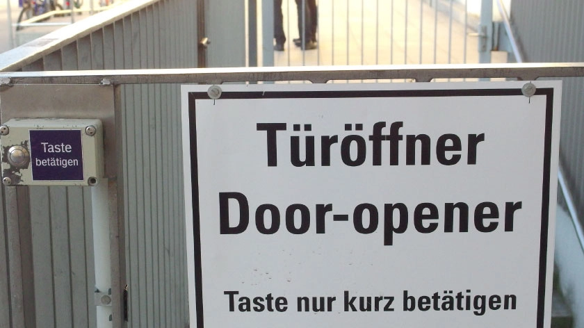 Hinweisschild an einem Tor: Türöffner, Door-opener, Taste nur kurz bestätigen. Neben dem Schild ist eine Taste angebracht.