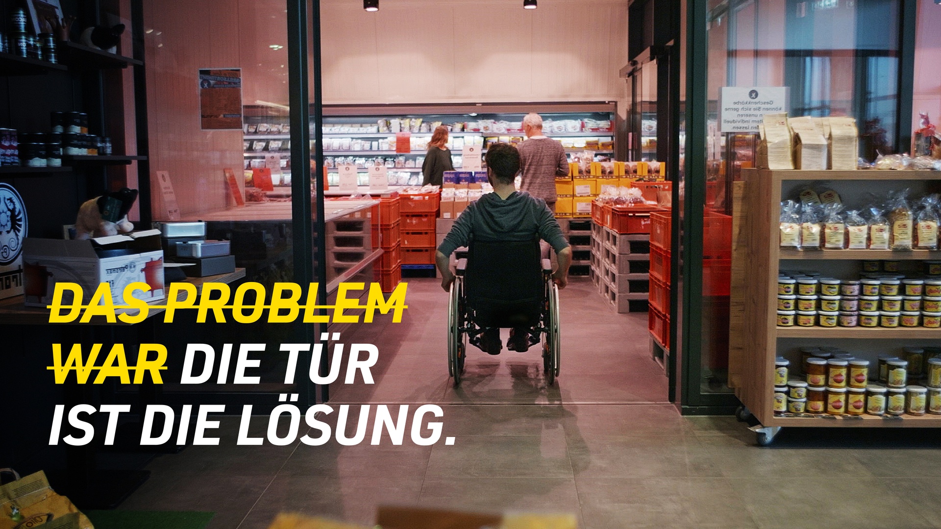 Ein Rollstuhlfahrer fährt durch eine schwellenlose Glasschiebetür in ein Geschäft. Auf dem Bild steht: „Die Tür ist die Lösung. Bauen Sie Barrieren ab – durch Zugangsmöglichkeiten für alle!“.